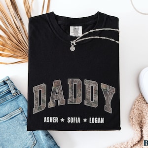 K&ouml;nnte beinhalten: Schwarzes T-Shirt mit dem Wort "DADDY" in einem Camouflage-Design. Darunter sind die Namen "ASHER", "SOFIA" und "LOGAN" aufgedruckt. Das Shirt wird mit blauen Jeans, wei&szlig;en Schuhen und einer Halskette gezeigt.