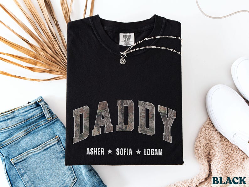 K&ouml;nnte beinhalten: Schwarzes T-Shirt mit dem Wort "DADDY" in einem Camouflage-Design. Darunter sind die Namen "ASHER", "SOFIA" und "LOGAN" aufgedruckt. Das Shirt wird mit blauen Jeans, wei&szlig;en Schuhen und einer Halskette gezeigt.