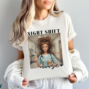 May include: Ivory-colored t-shirt featuring a graphic of a doll dressed as a nurse, with the words "NIGHT SHIFT" above the image. The word "nurse" is written in cursive below the doll. The shirt is being held by a person.