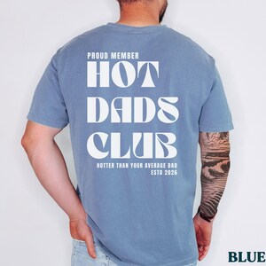 May include: Blue jean colored t-shirt with the text "PROUD MEMBER HOT DADS CLUB" in white block letters. The text "Hotter Than Your Average Dad ESTD 2026" is printed below. The shirt is a solid color.