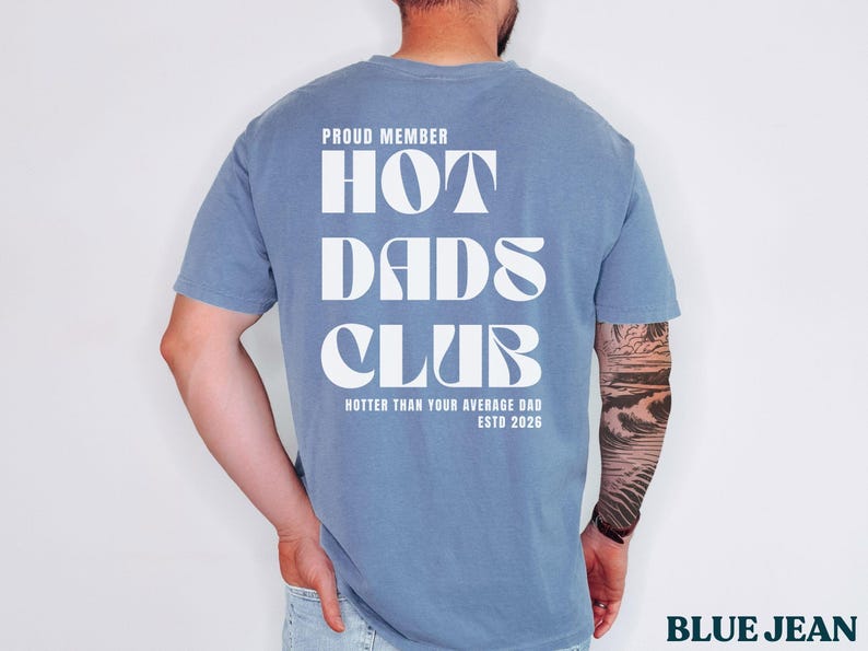 May include: Blue jean colored t-shirt with the text "PROUD MEMBER HOT DADS CLUB" in white block letters. The text "Hotter Than Your Average Dad ESTD 2026" is printed below. The shirt is a solid color.