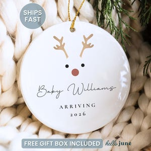 May include: White ceramic Christmas ornament with a reindeer face design. The ornament has brown antlers, black eyes, and a red nose. It is personalized with the name "Baby Williams" and the text "ARRIVING 2026". A gold string is attached for hanging.