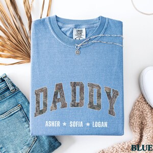 K&ouml;nnte beinhalten: Ein hellblaues Comfort Colors T-Shirt mit dem Wort "DADDY" in einem Camouflage-Design. Darunter stehen die Namen "Asher", "Sofia" und "Logan". Eine silberne Halskette und wei&szlig;e Turnschuhe sind ebenfalls im Bild.