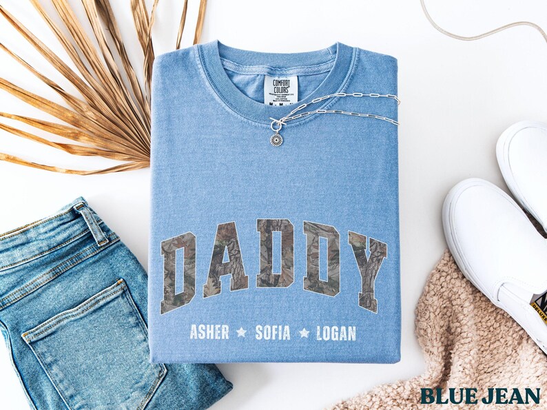 K&ouml;nnte beinhalten: Ein hellblaues Comfort Colors T-Shirt mit dem Wort "DADDY" in einem Camouflage-Design. Darunter stehen die Namen "Asher", "Sofia" und "Logan". Eine silberne Halskette und wei&szlig;e Turnschuhe sind ebenfalls im Bild.