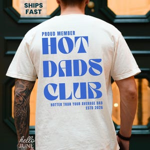 May include: Ivory-colored t-shirt with the text "PROUD MEMBER HOT DADS CLUB" in blue. The shirt also includes the text "HOTTER THAN YOUR AVERAGE DAD ESTD 2026". The shirt is a casual wear item.