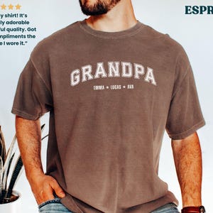 May include: Espresso-colored t-shirt with the word "GRANDPA" in white arched lettering. Below the word "GRANDPA" are the names "EMMA * LUCAS * AVA". The shirt has a vintage, worn look.