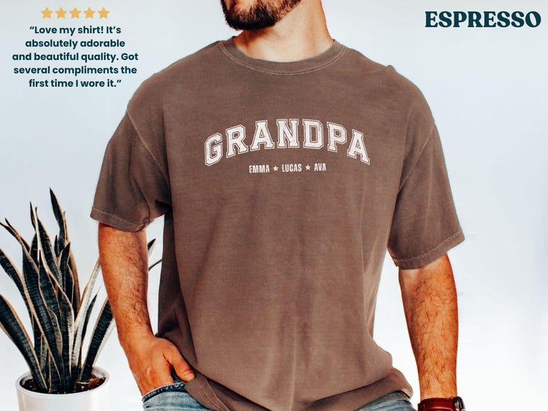 May include: Espresso-colored t-shirt with the word "GRANDPA" in white arched lettering. Below the word "GRANDPA" are the names "EMMA * LUCAS * AVA". The shirt has a vintage, worn look.
