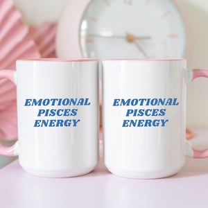 Op de afbeelding: Twee witte keramische mokken met roze randen. Beide mokken hebben de tekst "Emotional Pisces Energy" in blauw op de voorkant.