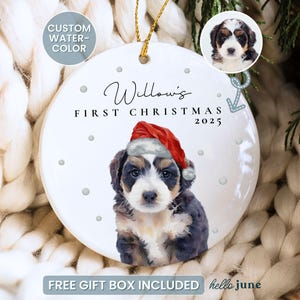 May include: A white ceramic Christmas ornament with a watercolor image of a puppy wearing a Santa hat. The ornament reads "Willow's First Christmas 2025" in script. The ornament has a gold string for hanging.