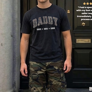 K&ouml;nnte beinhalten: Schwarzes T-Shirt mit dem Wort "DADDY" in einer verblassten Schriftart und den Namen "ASHER SOFIA LOGAN" darunter. Die Person tr&auml;gt eine Camouflage-Cargohose. Das Wort "BLACK" steht unten.