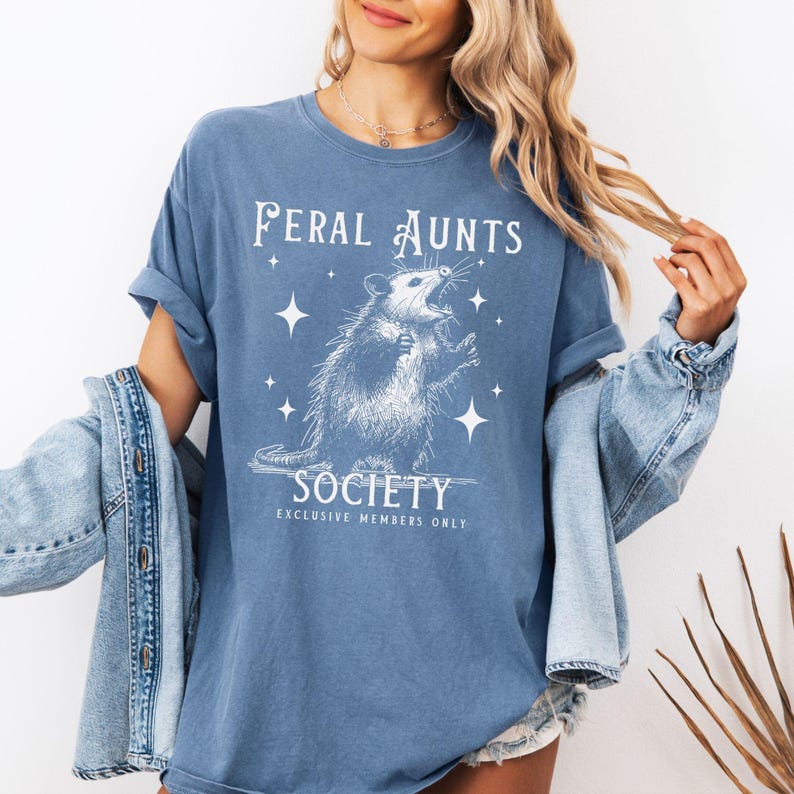 May include: A blue t-shirt with a white graphic of an opossum with stars and the text "Feral Aunts Society Exclusive Members Only".