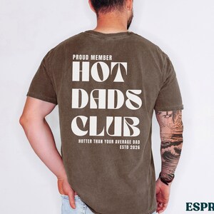 May include: Espresso-colored t-shirt with the text "PROUD MEMBER HOT DADS CLUB" in a bold, white font. The shirt also includes the phrase "HOTTER THAN YOUR AVERAGE DAD ESTD 2026".
