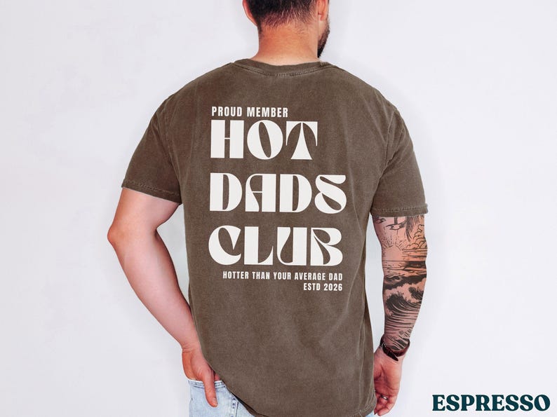 May include: Espresso-colored t-shirt with the text "PROUD MEMBER HOT DADS CLUB" in a bold, white font. The shirt also includes the phrase "HOTTER THAN YOUR AVERAGE DAD ESTD 2026".