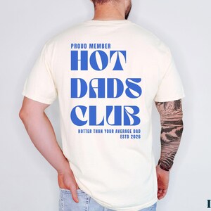 May include: Ivory-colored t-shirt with the text "PROUD MEMBER HOT DADS CLUB" in blue. The shirt also includes the phrase "HOTTER THAN YOUR AVERAGE DAD ESTD 2026". The shirt is a casual wear item.