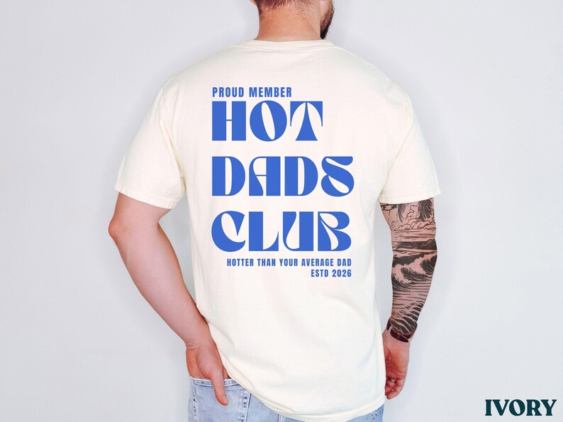 May include: Ivory-colored t-shirt with the text "PROUD MEMBER HOT DADS CLUB" in blue. The shirt also includes the phrase "HOTTER THAN YOUR AVERAGE DAD ESTD 2026". The shirt is a casual wear item.