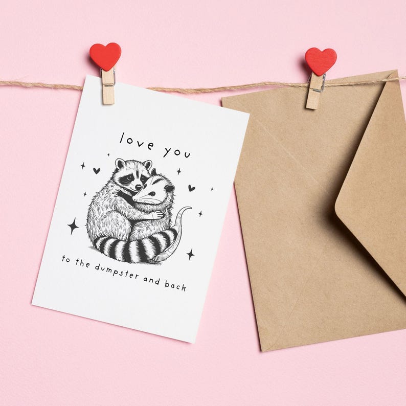 May include: A white card with a black and white illustration of a raccoon and opossum hugging. The card says "love you to the dumpster and back".