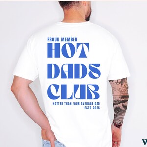May include: White t-shirt with the text "PROUD MEMBER HOT DADS CLUB" in blue, with "Hotter Than Your Average Dad ESTD 2026" below. The shirt is a classic crew neck style.
