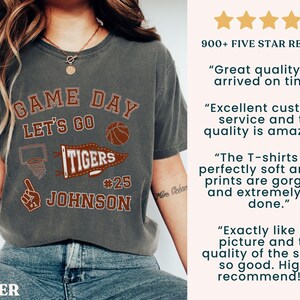 May include: Dark grey t-shirt with a basketball-themed graphic. The design includes the words "GAME DAY LET'S GO TIGERS #25 JOHNSON" in a distressed style. The shirt also features a basketball and a basketball hoop.