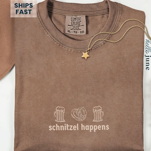 May include: Espresso-colored t-shirt with the phrase "schnitzel happens" and beer mugs and a pretzel graphic. A gold star pendant necklace rests on the shirt. The shirt is size XL-TG-EG.