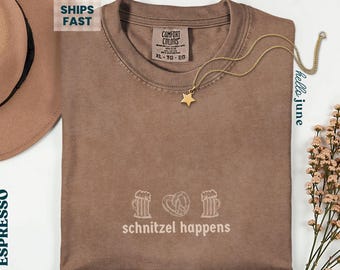May include: Espresso-colored t-shirt with the phrase "schnitzel happens" and beer mugs and a pretzel graphic. A gold star pendant necklace rests on the shirt. The shirt is size XL-TG-EG.