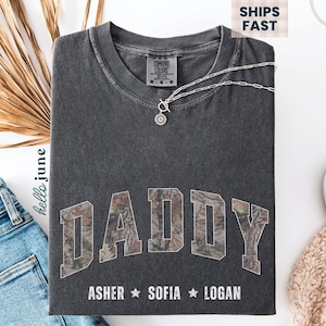 K&ouml;nnte beinhalten: Dunkelgraues T-Shirt mit dem Wort "DADDY" in einem Camouflage-Design. Darunter stehen die Namen Asher, Sofia und Logan. Eine silberne Halskette mit Anh&auml;nger liegt auf dem Shirt.