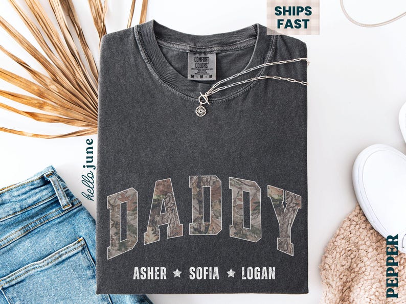 K&ouml;nnte beinhalten: Dunkelgraues T-Shirt mit dem Wort "DADDY" in einem Camouflage-Design. Darunter stehen die Namen Asher, Sofia und Logan. Eine silberne Halskette mit Anh&auml;nger liegt auf dem Shirt.