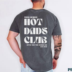 May include: Dark gray t-shirt with the text "PROUD MEMBER HOT DADS CLUB" in white, with "Hotter Than Your Average Dad ESTD 2026" below. The shirt is worn by a person.