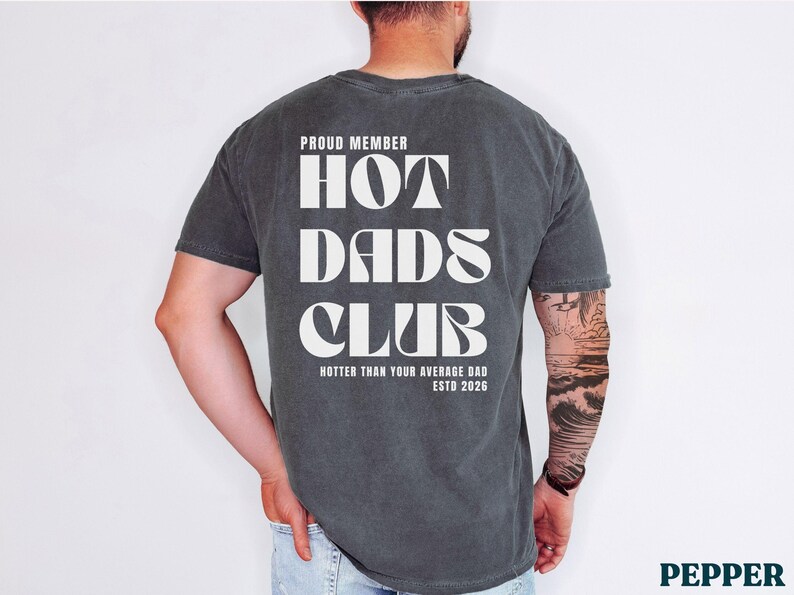 May include: Dark gray t-shirt with the text "PROUD MEMBER HOT DADS CLUB" in white, with "Hotter Than Your Average Dad ESTD 2026" below. The shirt is worn by a person.