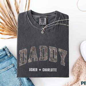 K&ouml;nnte beinhalten: Dunkelgraues T-Shirt mit dem Wort "DADDY" im Camouflage-Druck. Die Namen "Asher" und "Charlotte" sind darunter aufgedruckt. Eine silberne Halskette mit Anh&auml;nger liegt auf dem Shirt. Blaue Jeans und wei&szlig;e Schuhe sind ebenfalls im Bild.