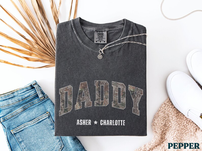 K&ouml;nnte beinhalten: Dunkelgraues T-Shirt mit dem Wort "DADDY" im Camouflage-Druck. Die Namen "Asher" und "Charlotte" sind darunter aufgedruckt. Eine silberne Halskette mit Anh&auml;nger liegt auf dem Shirt. Blaue Jeans und wei&szlig;e Schuhe sind ebenfalls im Bild.