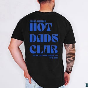 May include: Black t-shirt with the text "PROUD MEMBER HOT DADS CLUB" in blue. The shirt also includes the phrase "HOTTER THAN YOUR AVERAGE DAD ESTD 2026". The shirt is a classic crew neck.