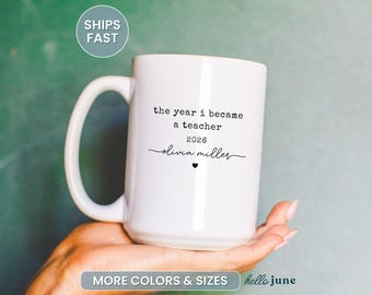 May include: A white ceramic mug with a large handle, featuring the text "the year i became a teacher 2026 olivia miller" in black. A small black heart is below the name. The mug is held in a hand against a green background.