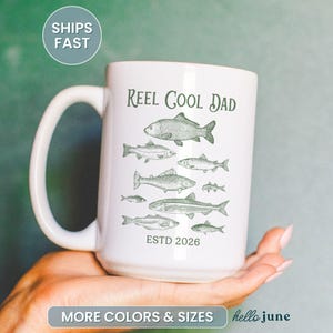 May include: White ceramic mug with a large handle, featuring the text "Reel Cool Dad" above a series of fish illustrations in green. The mug also includes the text "ESTD 2026".