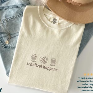 May include: Ivory-colored t-shirt with the phrase "schnitzel happens" and beer mugs and a pretzel graphic. The shirt is folded on top of blue jeans and a tan hat.