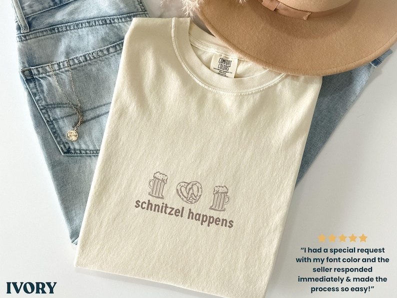 May include: Ivory-colored t-shirt with the phrase "schnitzel happens" and beer mugs and a pretzel graphic. The shirt is folded on top of blue jeans and a tan hat.