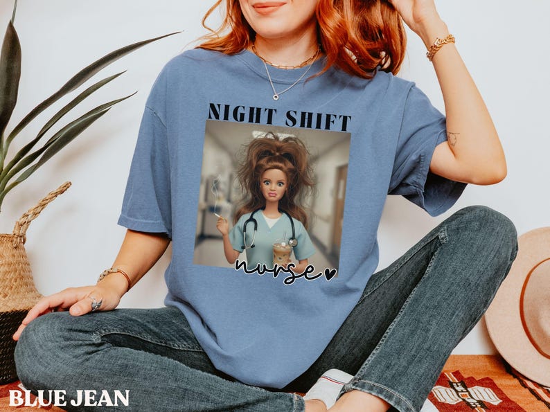 May include: A blue t-shirt with the words "NIGHT SHIFT" above an image of a doll dressed as a nurse. The doll is wearing a stethoscope and holding a coffee cup. The word "nurse" is written below the image.