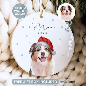 May include: A white ceramic Christmas ornament featuring a watercolor portrait of a dog wearing a Santa hat. The ornament has the name "Max" and the year "2025" written on it. The ornament is hanging by a gold string.