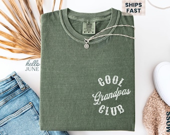 May include: An olive green t-shirt with the text "Cool Grandpas Club" in white script. A silver necklace with a pendant is draped across the shirt. The words "Ships Fast" are in the upper right corner.