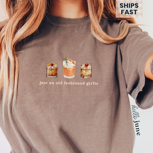 Może przedstawiać: Brązowy t-shirt z trzema ilustracjami koktajli i napisem "just an old fashioned girlie". Koszulka ma luźny kr&oacute;j i kr&oacute;tkie rękawy. Kolor przypomina espresso.