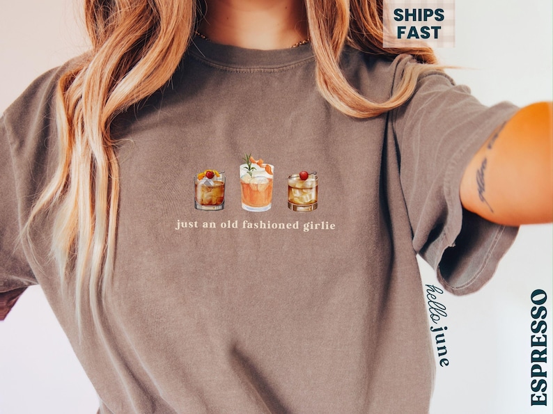 Może przedstawiać: Brązowy t-shirt z trzema ilustracjami koktajli i napisem "just an old fashioned girlie". Koszulka ma luźny kr&oacute;j i kr&oacute;tkie rękawy. Kolor przypomina espresso.