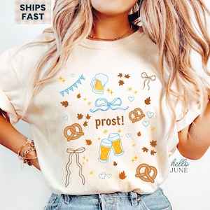 May include: Ivory-colored t-shirt with a festive design featuring pretzels, beer steins, and the word "prost!" in brown. The design includes blue and brown accents, perfect for Oktoberfest celebrations.