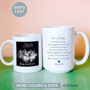 May include: Two white ceramic mugs. One mug features a black and white sonogram image with the text "Baby Smith." The other mug has the text "Dear Daddy" and a poem about Father's Day. The mugs are on a green surface.