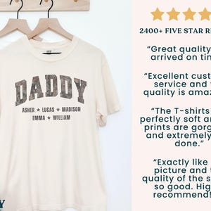 K&ouml;nnte beinhalten: Cremefarbenes T-Shirt mit dem Wort "DADDY" in Camouflage-Schrift. Darunter stehen die Namen Asher, Lucas, Madison, Emma und William. Das T-Shirt h&auml;ngt an einem Holzkleiderb&uuml;gel.