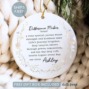 May include: A white ceramic ornament with the text "Difference Maker" and a definition. The ornament has a floral design and a gold string for hanging. The text "Ashley" is also included.