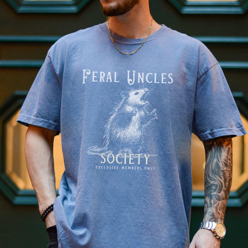 May include: A light blue t-shirt with a white graphic of a possum with the text "Feral Uncles Society Exclusive Members Only".