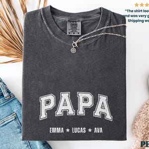 May include: Dark gray t-shirt with the word "PAPA" in white, collegiate-style lettering. Below the word "PAPA" are the names EMMA, LUCAS, and AVA. A silver necklace rests on the shirt. The shirt is folded on a white surface.