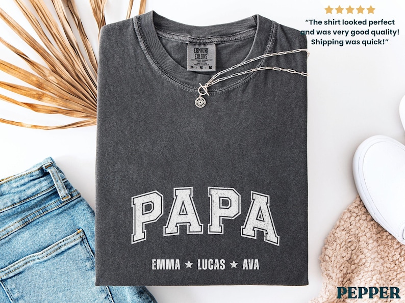 May include: Dark gray t-shirt with the word "PAPA" in white, collegiate-style lettering. Below the word "PAPA" are the names EMMA, LUCAS, and AVA. A silver necklace rests on the shirt. The shirt is folded on a white surface.