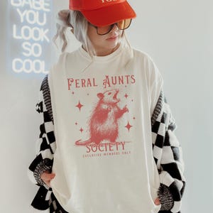 May include: A white long-sleeved shirt with a red graphic of a possum with the text "Feral Aunts Society Exclusive Members Only".