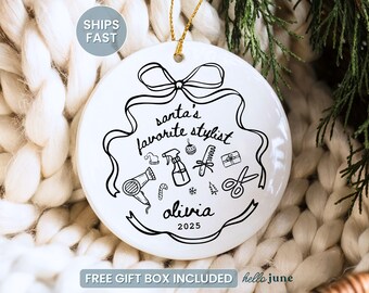 May include: White ceramic Christmas ornament with black line art. The ornament features the text "Santa's Favorite Stylist" with illustrations of salon tools. The name "Olivia" and the year "2025" are also included.