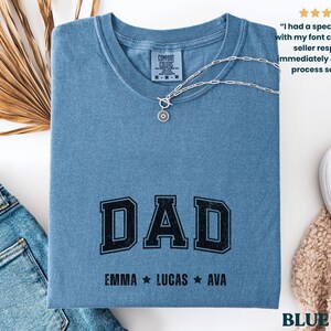 May include: A blue jean-colored t-shirt with the word "DAD" in black block letters. Below the word "DAD" are the names "EMMA", "LUCAS", and "AVA". A silver necklace with a pendant rests on the shirt. The shirt is folded and placed on a white surface.